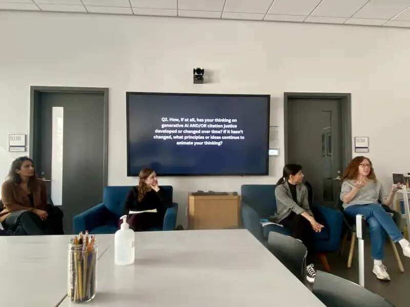 Four women sit in chairs in the DHC, one is holding a microphone and speaking. In the background is a monitor displaying a question.