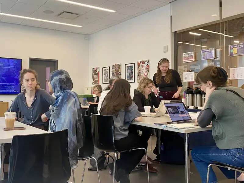 Faculty sit in groups in the Digital Humanities Center, with laptops out. Elana is standing up, consulting with a faculty member.